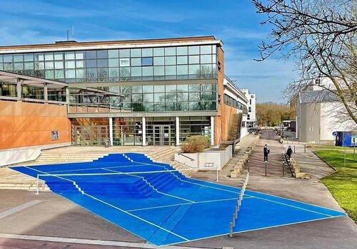 Parvis de la Maison de l'Université de Rouen Normandie, recouvert de l'oeuvre de Laurent Perbos, un court de tennis grandeur nature bleu aux bandes blanches.