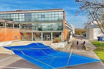 Parvis de la Maison de l'Université de Rouen Normandie, recouvert de l'oeuvre de Laurent Perbos, un court de tennis grandeur nature bleu aux bandes blanches.